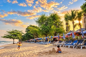 A beach with rows of sun loungers and umbrellas, a child playing with sand, and palm trees. - Near Beach Condo With Pool Access in Pattaya City (Pattaya)