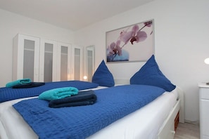 A hotel room with two beds, each with a blue bedspread and pillows, a white bedside table, and a wall-mounted picture of flowers. - Holiday apartment with 2 bedrooms (Borkum)