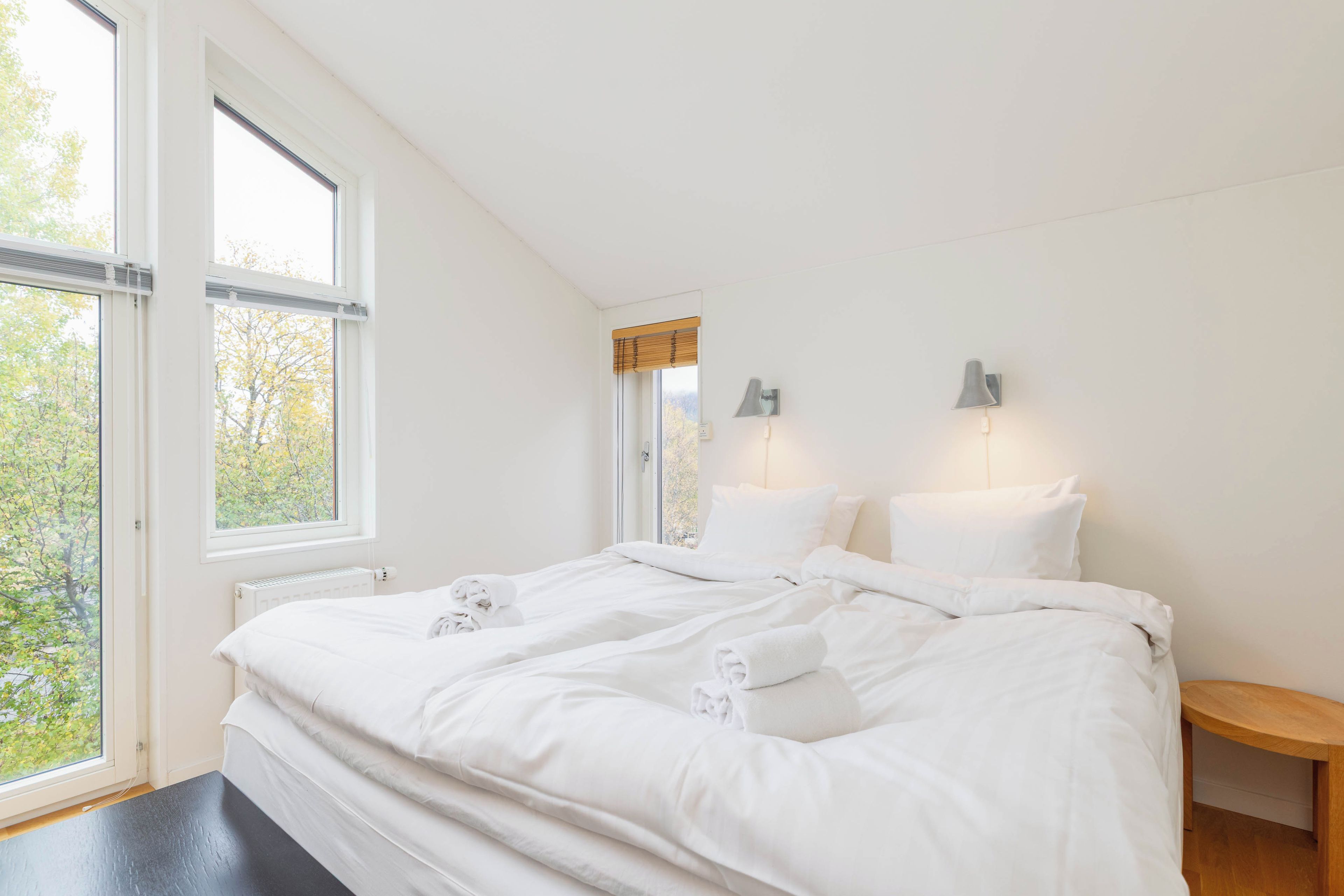 A bedroom with a large window, a bed with white linens, a bedside table, and two wall-mounted lamps.