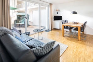 A modern living room with a sofa, a dining table with chairs, a TV, and a sliding glass door leading to an outdoor area with a chair and umbrella. - Norderney Sun Decks Apt. 11 (Norderney)