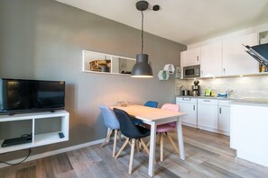 A modern kitchen with white cabinets, a dining table with chairs, a hanging pendant light, and a built-in TV shelf. - Holiday apartment with 1 bedroom (Borkum)