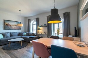 A modern living room with a sofa, dining table, and chairs. There is a large window with curtains, a hanging lamp, and a painting on the wall. - Holiday apartment with 1 bedroom (Borkum)