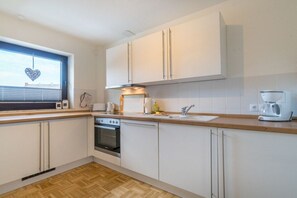 A modern kitchen with white cabinets, a built-in oven, a coffee maker, and a window with blinds. - Holiday apartment Sonneninsel (Borkum)
