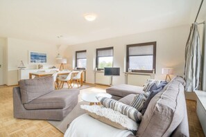 A modern living room with a grey sofa set, a dining area with a table and chairs, and a kitchen area with a TV. - Holiday apartment Sonneninsel (Borkum)