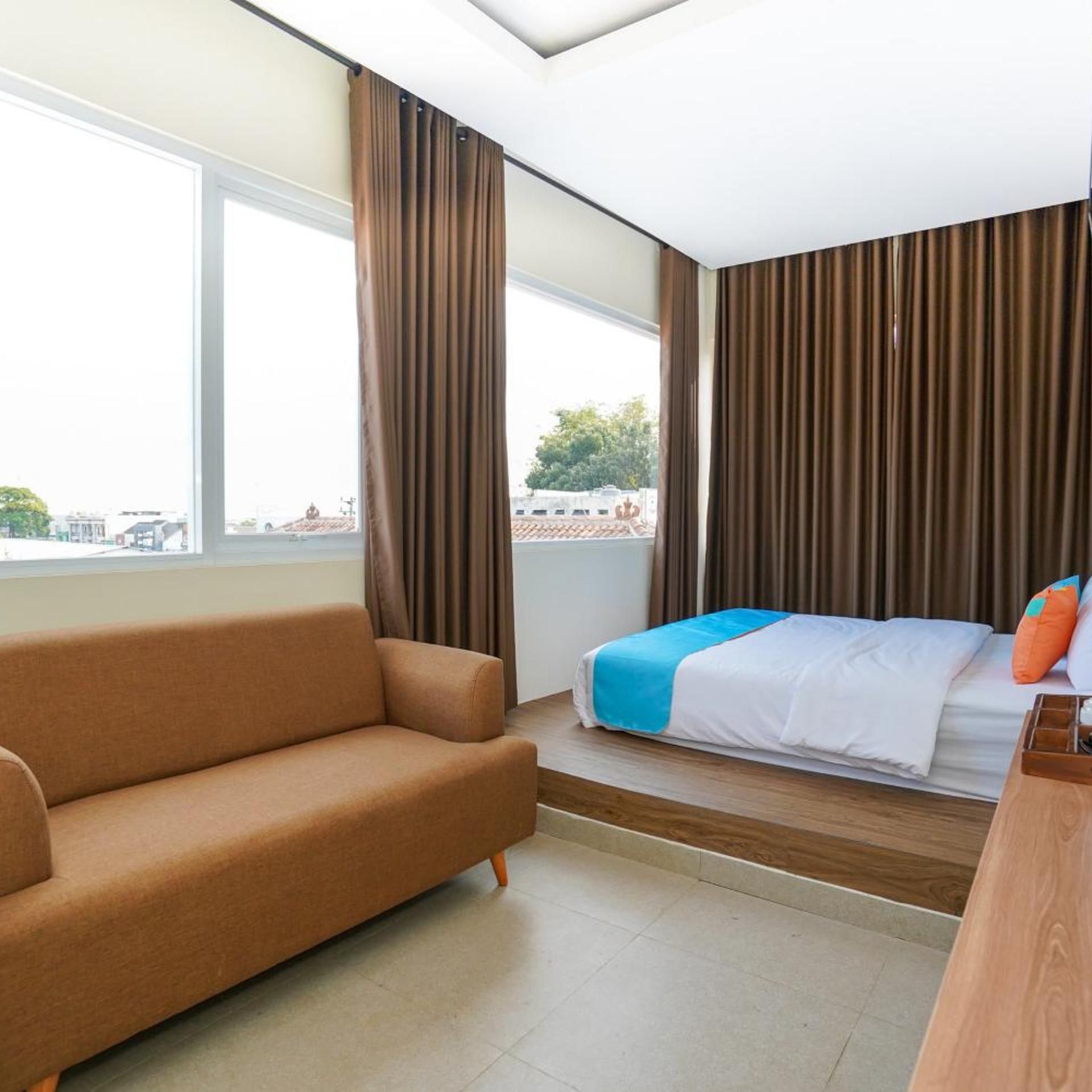 A hotel room with a bed, a sofa, a window with curtains, and a view of buildings outside.