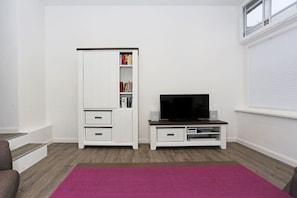 Une pièce avec une armoire blanche, un téléviseur posé sur un meuble en bois et une fenêtre avec des stores.