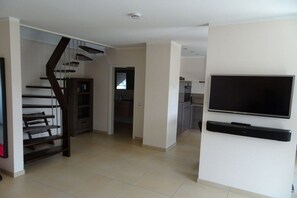 A modern living room with a flat-screen TV mounted on a white wall, a staircase with dark railings, and a kitchen area in the background. - Holiday apartment with 2 bedrooms (Borkum)