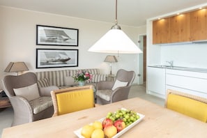 Ett modernt vardagsrum med ett matbord, två fåtöljer, en soffa och en köksdel med träskåp och en hängande lampa.