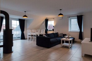 A modern living room with a sofa, a coffee table, and a TV stand. There is a dining area with chairs and a table. The room has large windows with curtains. - Holiday apartment with 2 bedrooms (Krummhörn)