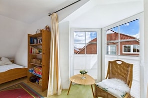 A bedroom with a bed, a bookshelf, a small table with a vase, a wicker chair, and a window with a view of buildings. - Holiday home in Dornum with Wi-Fi and gym (Dornum)