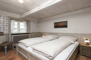 A bedroom with a double bed, a window with curtains, a radiator, a picture on the wall, and a bedside table with a lamp. - Old Beach Warden's Office (List)