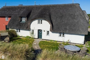 A thatched-roof house with a white exterior, a circular door, and a small porch with wicker furniture. - Old Beach Warden's Office (List)