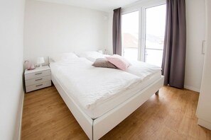 A bedroom with a white bed, a nightstand with a lamp, and a window with curtains. - Holiday apartment with 3 bedrooms (Norderney)