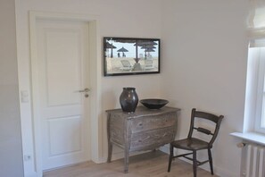 Een kamer met een witte deur, een vintage dressoir met een vaas en schaal, een houten stoel en een lijst met daarop een afbeelding van een strand.