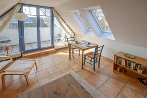 A cozy room with a dining table, chairs, a bookshelf, and a large window with curtains. - Sonnenhof Top Floor D5 (Prerow)
