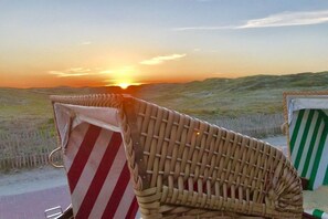 A beach chair with a striped towel and a woven beach chair, both positioned on a sandy area with a view of the sunset over dunes. - Holiday apartment with 1 bedroom (Norderney)