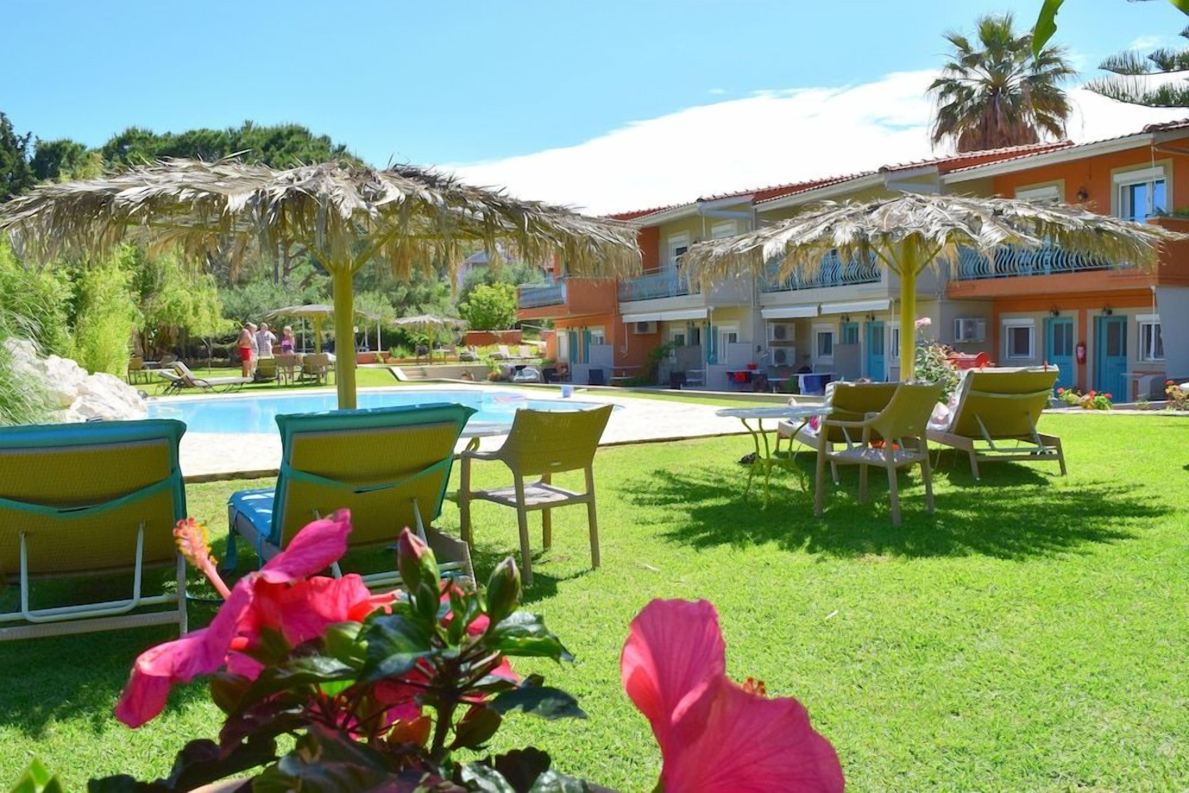A pool area with lounge chairs and umbrellas, a building with balconies, and a well-maintained lawn.