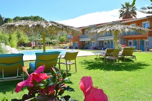 Zona de piscina con sillones de descanso y sombrillas, un edificio con balcones y un césped bien cuidado.