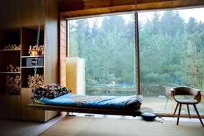 Un dormitorio con una cama colgante, una silla y vistas a un bosque a través de un ventanal grande.