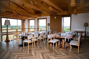 Un comedor con varias mesas y sillas, amplias ventanas con vistas al exterior y vigas de madera en el techo.