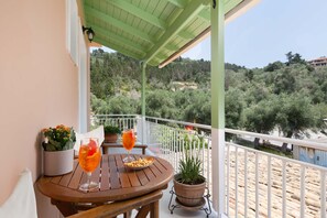 A wooden table with two glasses of orange beverage, a plate with snacks, and potted plants on a balcony with a view of a forested hillside. - Venti Di Paxos 2 | One -Bedroom Apartment, Apartment (Paxoi)