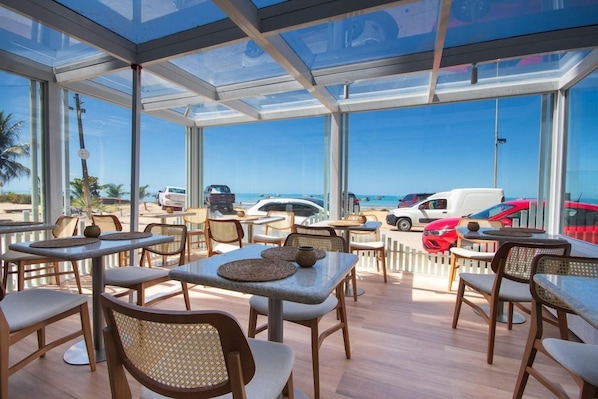 An outdoor seating area with tables and chairs, large windows, and a view of the beach and parked cars. - Janga Boutique Hotel (Maragogi)