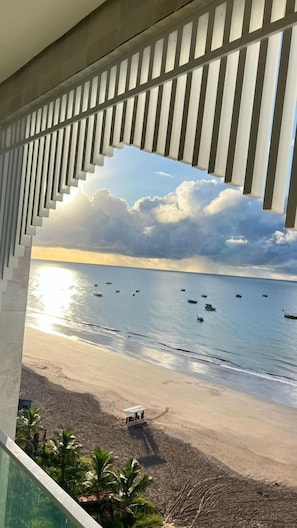 Blick auf einen Strand durch ein Fenster mit einem gemusterten Rollo, Boote auf dem Wasser und ein Strandtuch.