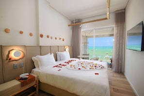 A hotel room with a large bed, a bedside table with a phone and remote, a wall-mounted TV, and a view of the ocean through large windows. - Janga Boutique Hotel (Maragogi)