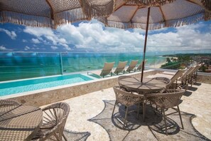 Ein Poolbereich mit Liegestühlen und einem Tisch, mit Blick auf ein klares blaues Meer und einen fernen Strand.