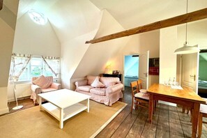 A cozy loft bedroom with a sloped ceiling, a wooden beam, a white bed, a small table, and a window with curtains. - Old Registry Office Large Apartment (Oldsum)