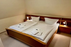 A double bed with white linens and two pillows, a wooden headboard, bedside tables with lamps, and a carpeted floor. - Hansen Grevelingstieg 16 (Nieblum)