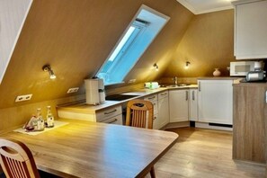 A kitchen with a sloped ceiling, a window, white cabinets, a wooden table, and a microwave. - Hansen Grevelingstieg 16 (Nieblum)