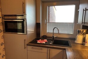 A compact kitchen with a built-in oven, a window with blinds, a sink, and a coffee maker. - Holiday apartment in Wyk on Föhr with balcony (Wyk auf Föhr)