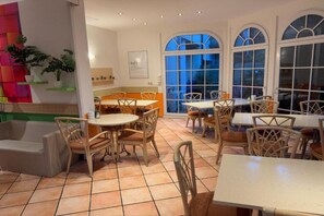 A dining area with multiple tables and chairs, a kitchen in the background, and large windows. - Sunday Hotel Leipzig Südvorstadt (Markgraf) - Standard Single Room (Leipzig)