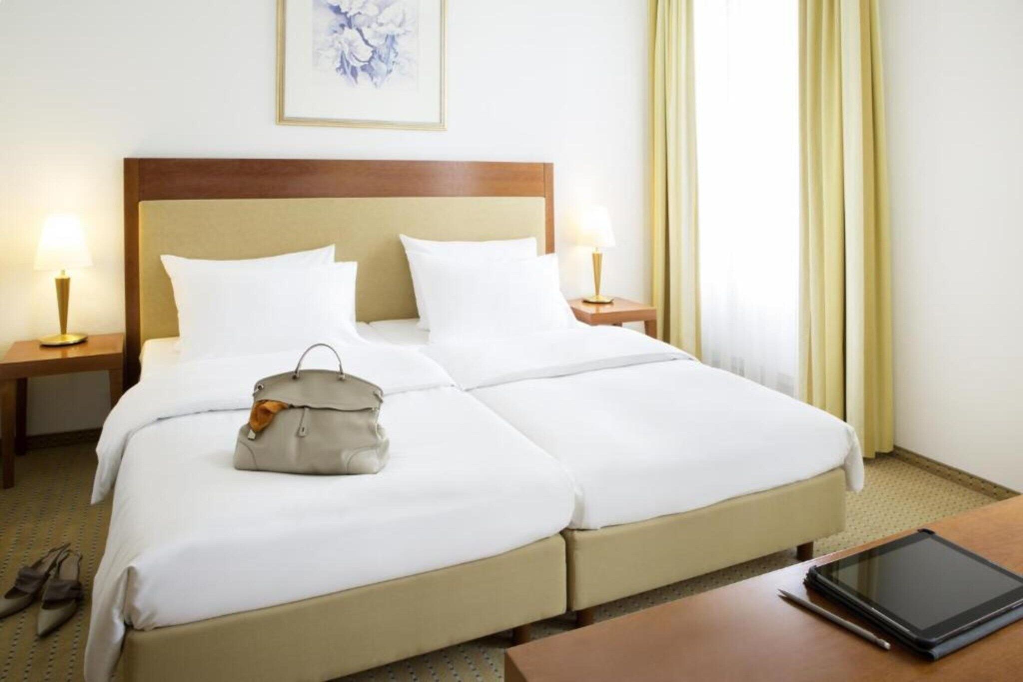 Habitación de hotel con cama doble, dos mesitas de noche, una lámpara, un bolso de mano y una tableta sobre una mesa.