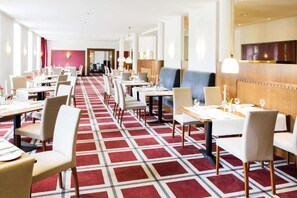 Zona de menjador d'hotel amb diverses taules i cadires, una catifa estampada i un disseny interior modern.