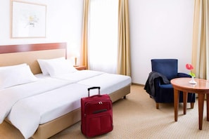 Una habitación de hotel con una cama, una maleta roja, un sillón azul, una mesita redonda y un cuadro colgado en la pared.