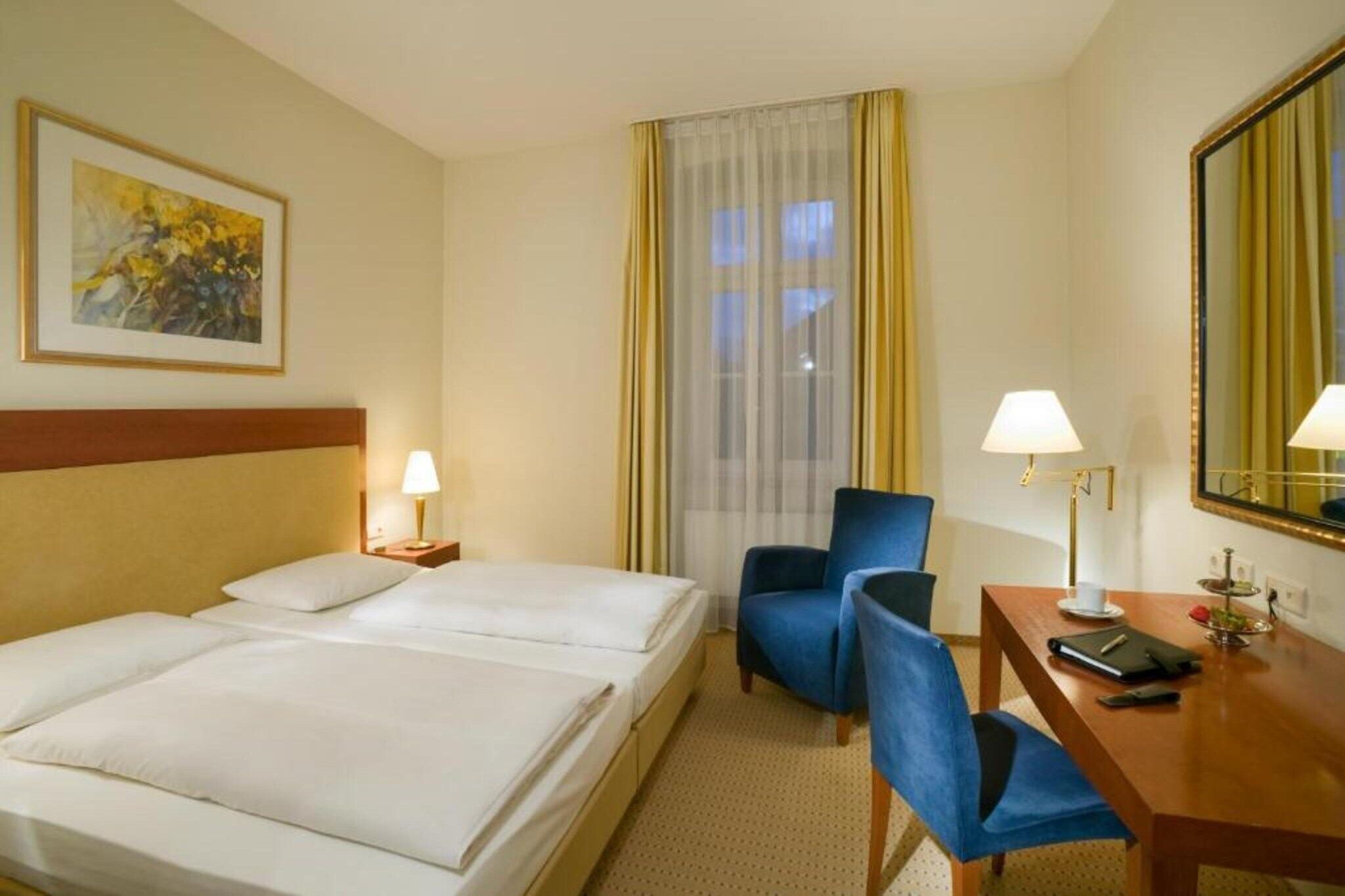 Habitació d'hotel amb dos llits, un escriptori amb cadira, una làmpada, un mirall i una finestra amb cortines.