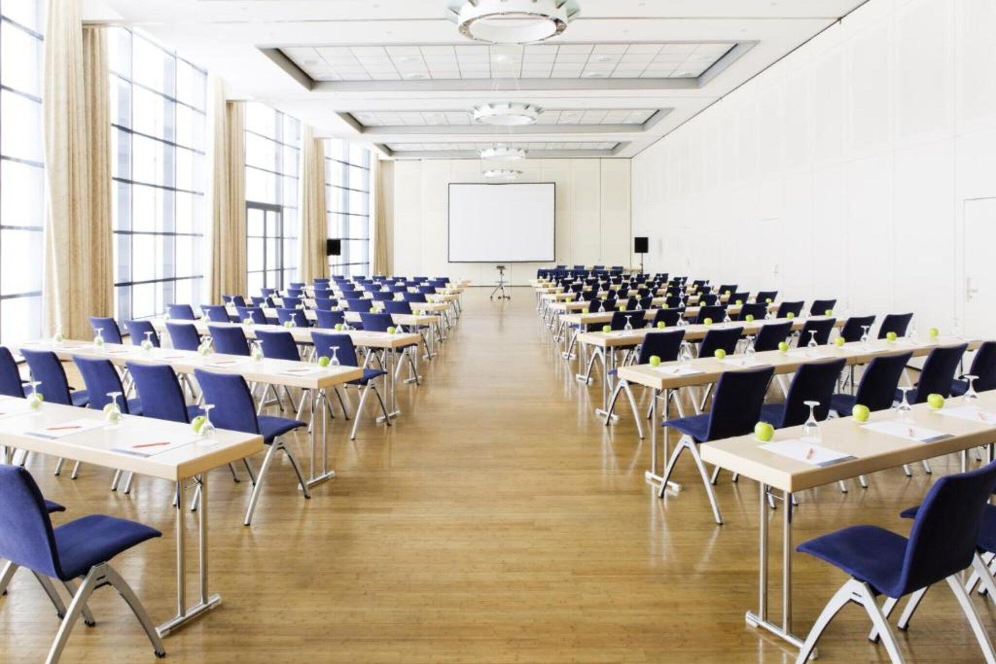 Un salón de conferencias con filas de mesas y sillas, una pantalla de proyector y ventanas amplias.