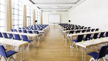 Un salón de conferencias con filas de mesas y sillas, una pantalla de proyector y ventanas amplias.