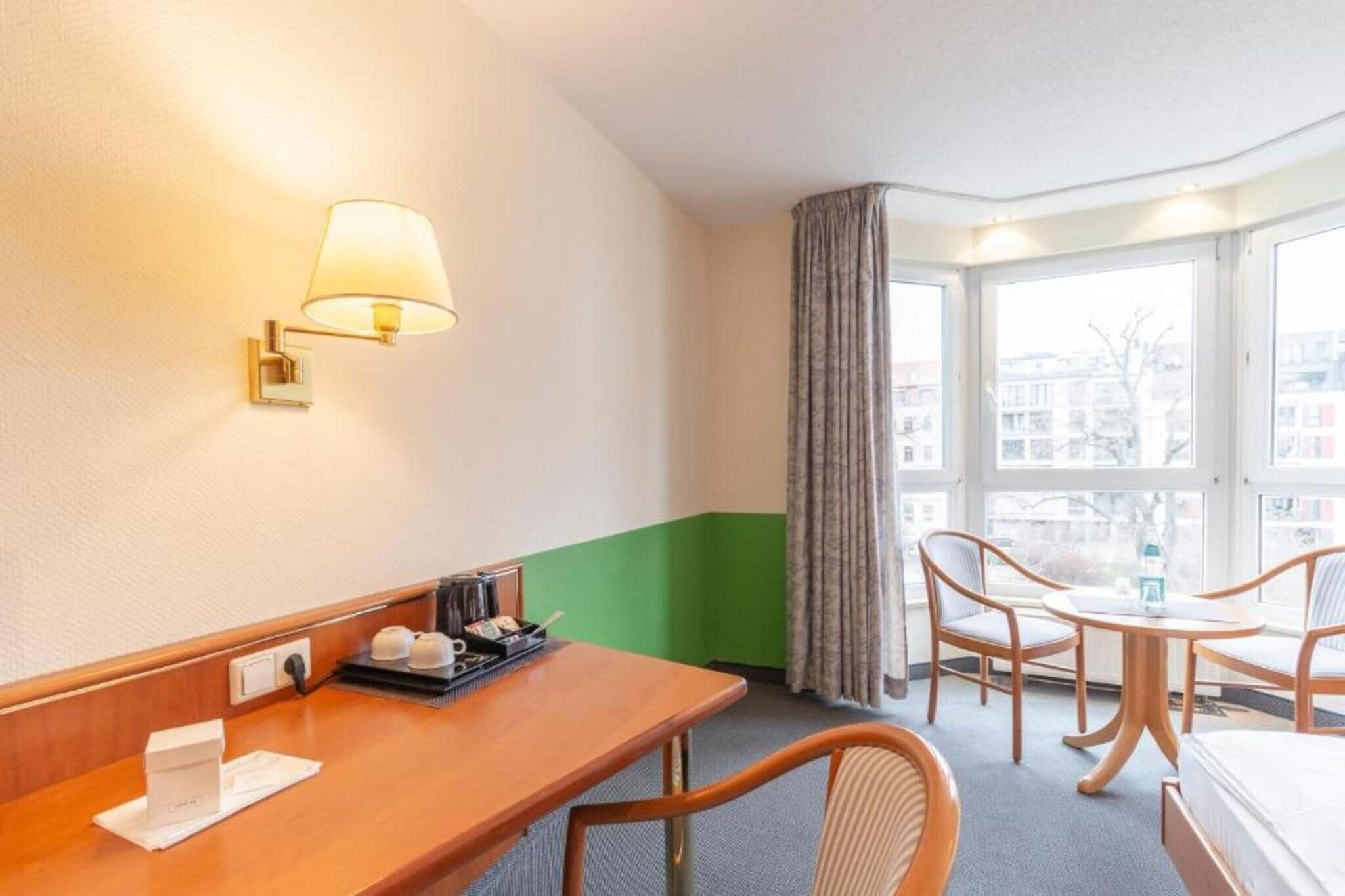 Ein Hotelzimmer mit einem Holztisch, einem Stuhl, einem kleinen Tisch, einer Lampe, einem Fenster mit Vorhängen und Blick auf Gebäude.