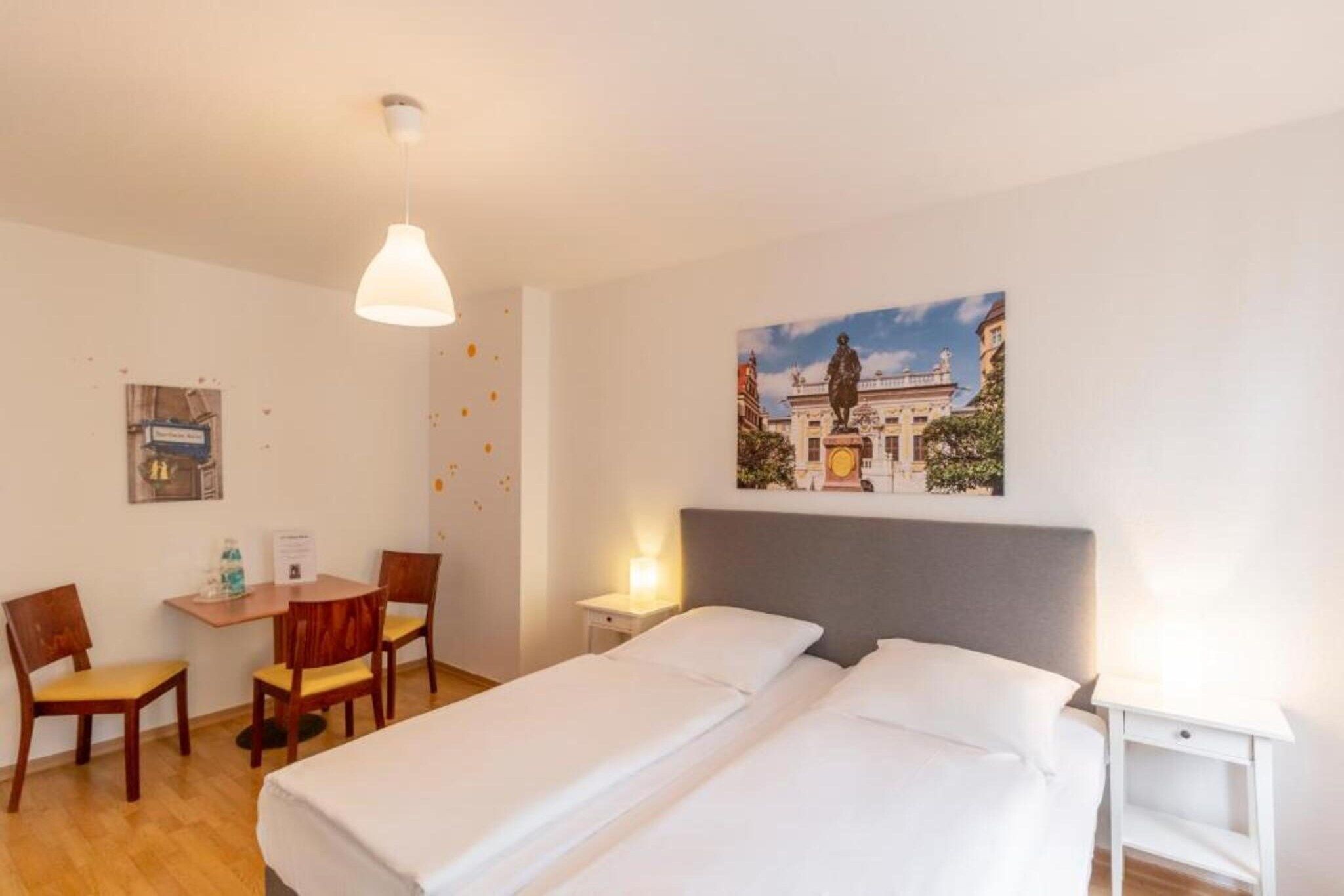 A hotel room with two beds, a desk with chairs, a painting of a building, and a ceiling light.