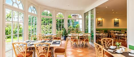 A spacious indoor dining area with multiple tables and chairs, large windows offering views of greenery, and a ceiling with recessed lighting.