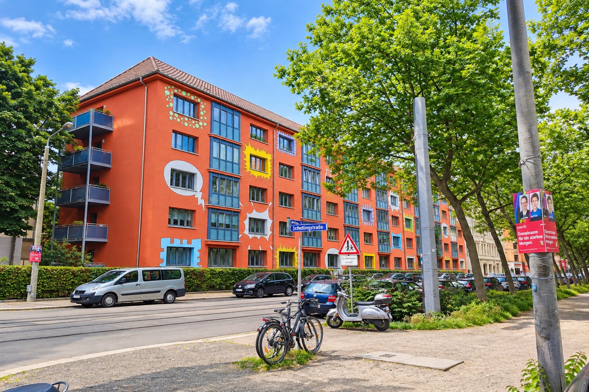 A multi-story building with balconies, a street sign, parked cars, and a pedestrian crossing sign.