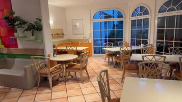 Un comedor con varias mesas y sillas, una cocina al fondo y ventanas amplias.