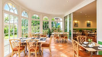 A spacious indoor dining area with multiple tables and chairs, large windows offering views of greenery, and a ceiling with recessed lighting.