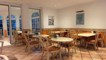 A restaurant interior with round tables and wicker chairs, a tiled floor, and a poster on the wall.