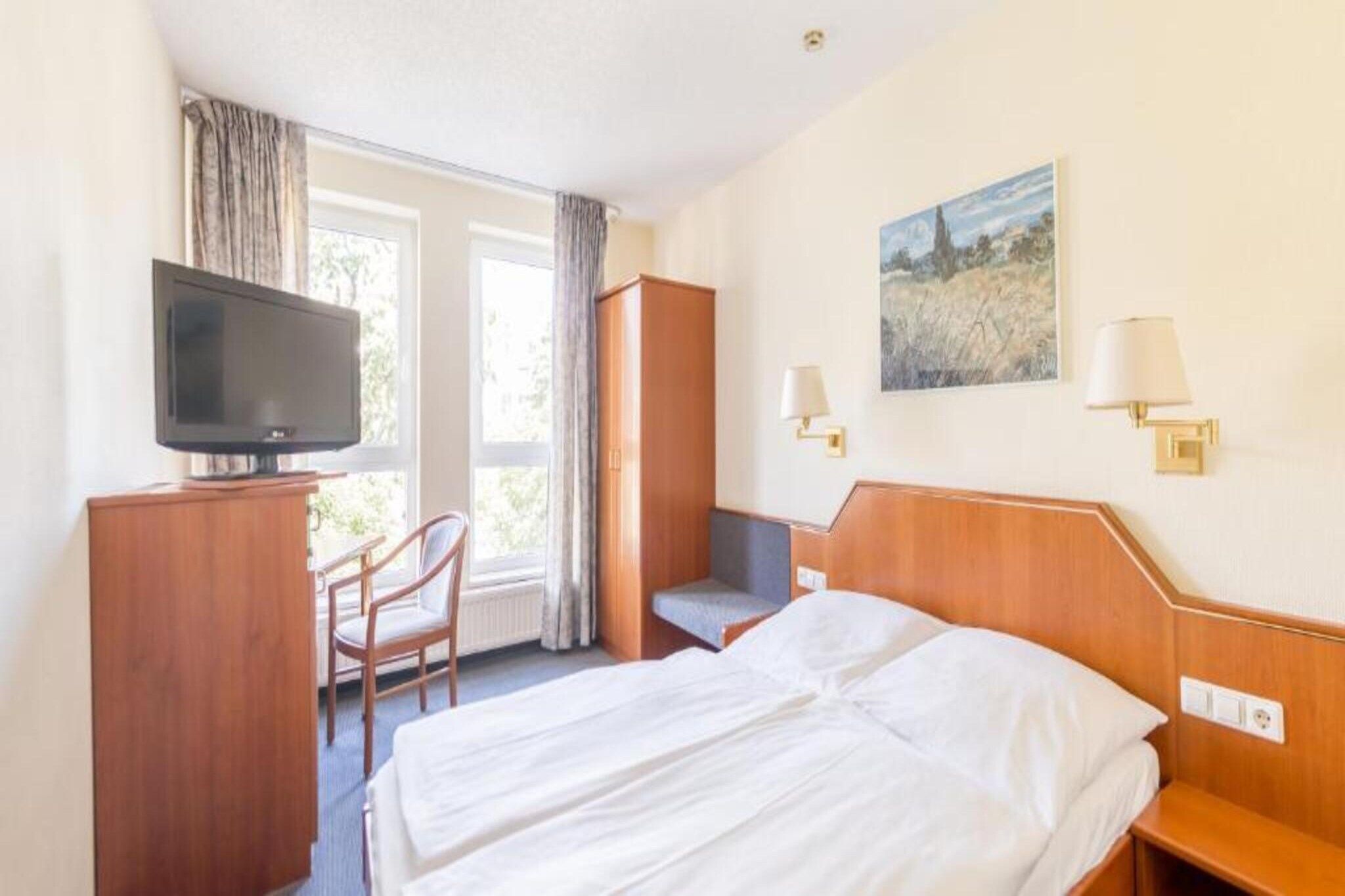 A hotel room with a bed, a desk with a television, a chair, a small table, and a painting on the wall.