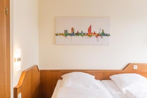 Habitación de hotel con cama, cabecera de madera y un cuadro en la pared que muestra el horizonte de una ciudad.