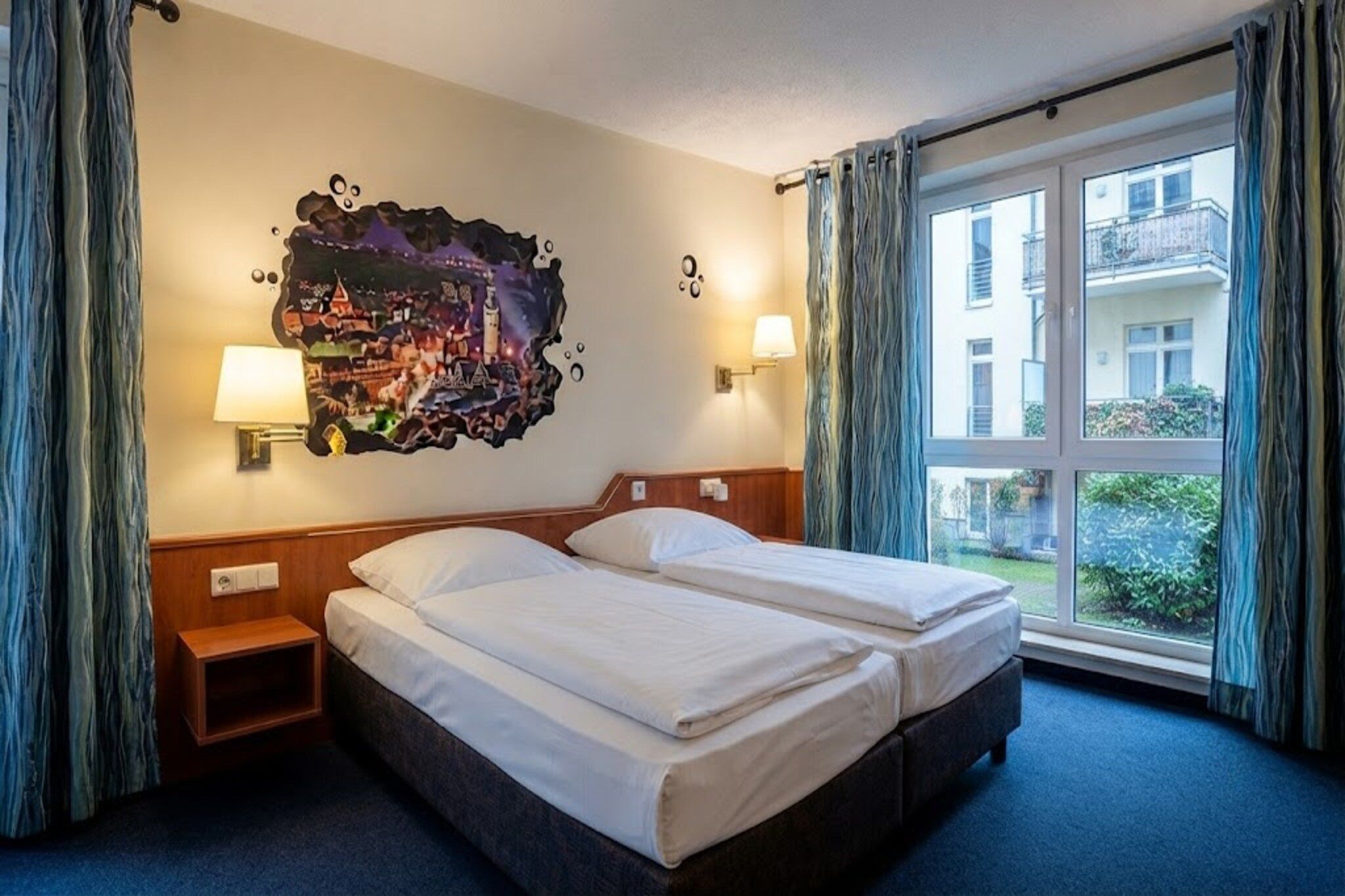 A hotel room with a double bed, two bedside lamps, a wall mural, and a large window with curtains.
